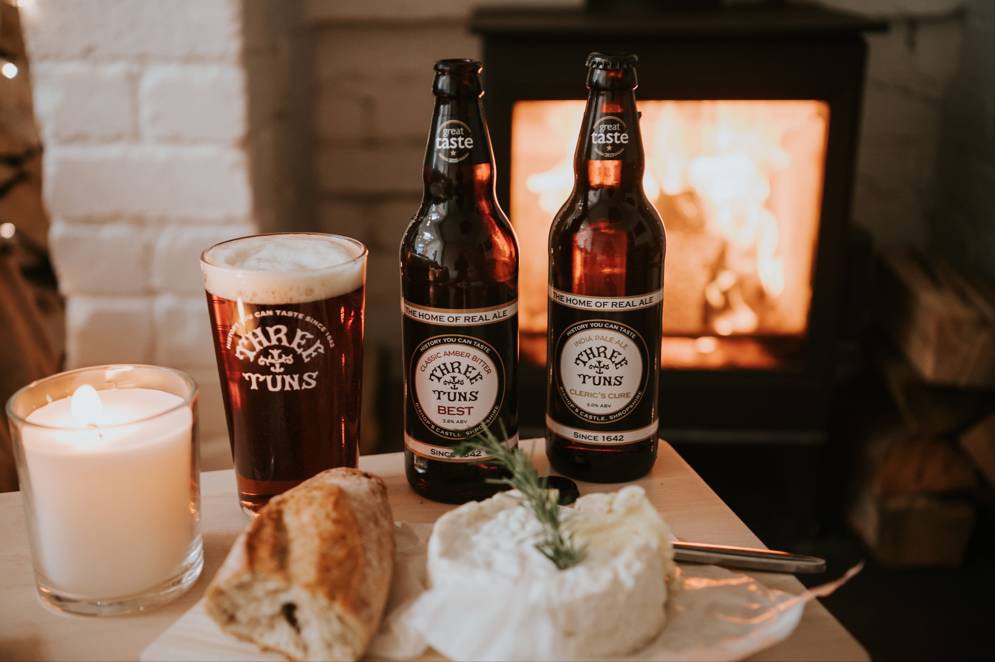 two bottles and pint glass of three tuns brewery beer on a table with a candle and bread in front of a lit log burner fire
