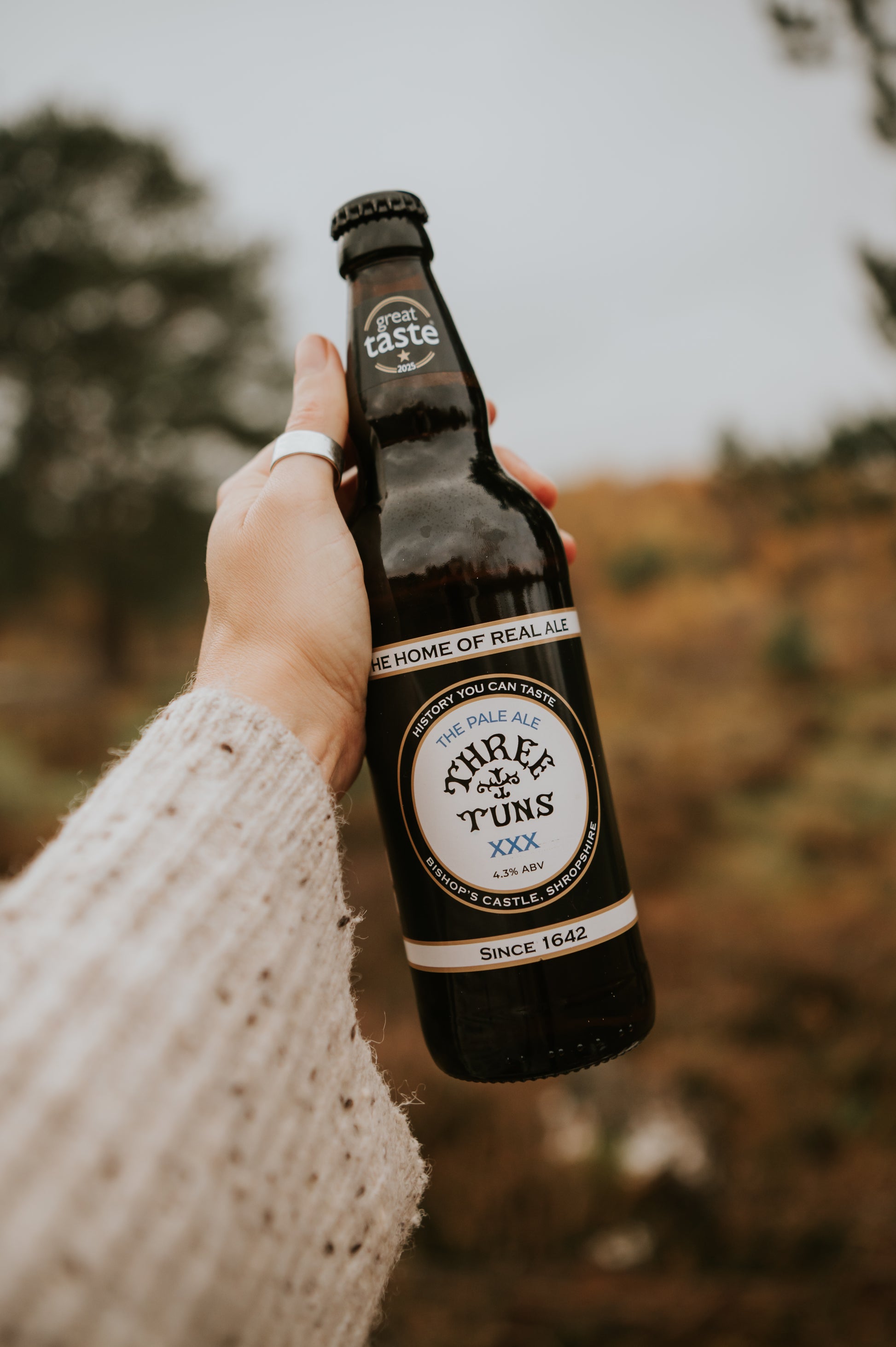 hand holding a bottle of three tuns brewery xxx pale ale beer with a blurred natural countryside background